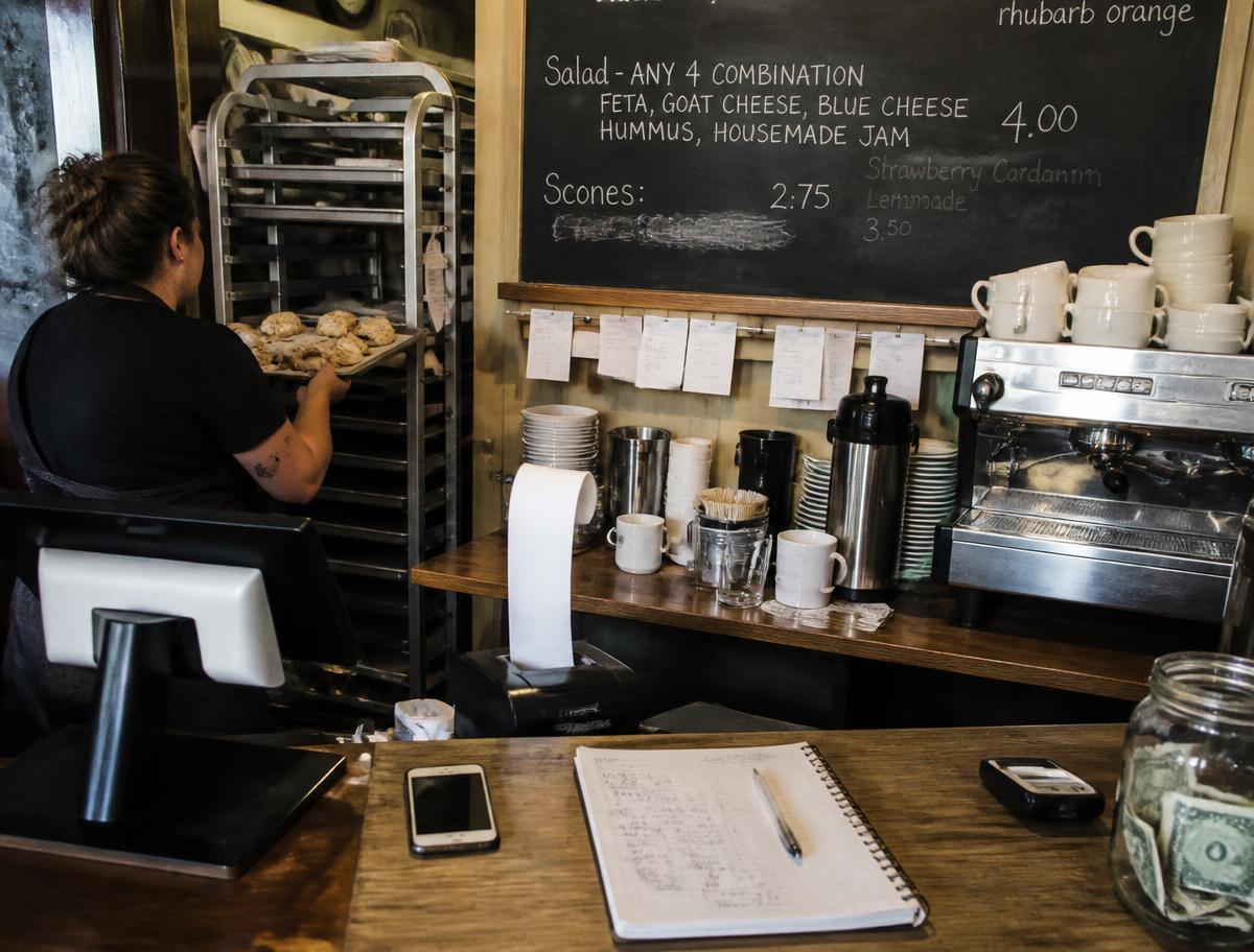 Behind the counter of a small bakery cafe: a tray of scones, chalkboard menu, order tickets on a rail, and a notebook of numbers by the register